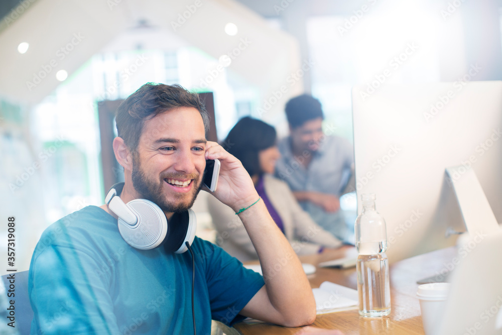 Fototapeta premium Smiling creative businessman with headphones talking on cell phone in office