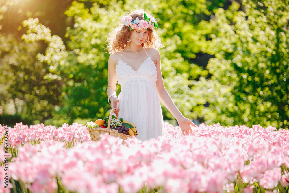 Young happy girl in a tulip field. Smiling curly woman with a basket of fruit in a sunny flowering meadow. Positive model with a wreath of flowers on her hair. ..Summer scene. Young happy girl in a tulip field. Smiling curly woman with a basket of fruit in a sunny flowering meadow. Positive model with a wreath of flowers on her hair. ..Summer scene.
