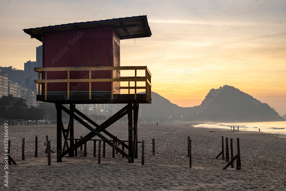 Old red and yellow wooden vintage lifeguard tower on the beach of Copacabana in Rio de Janeiro backlit by intense golden hour sunrise sunlight with Leme mountain in the background