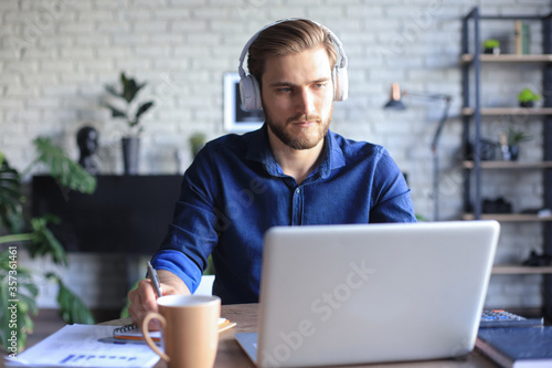 Quadro em tela Confident businessman in earphones is writing notes or financial report while sitting at desk with laptop at home