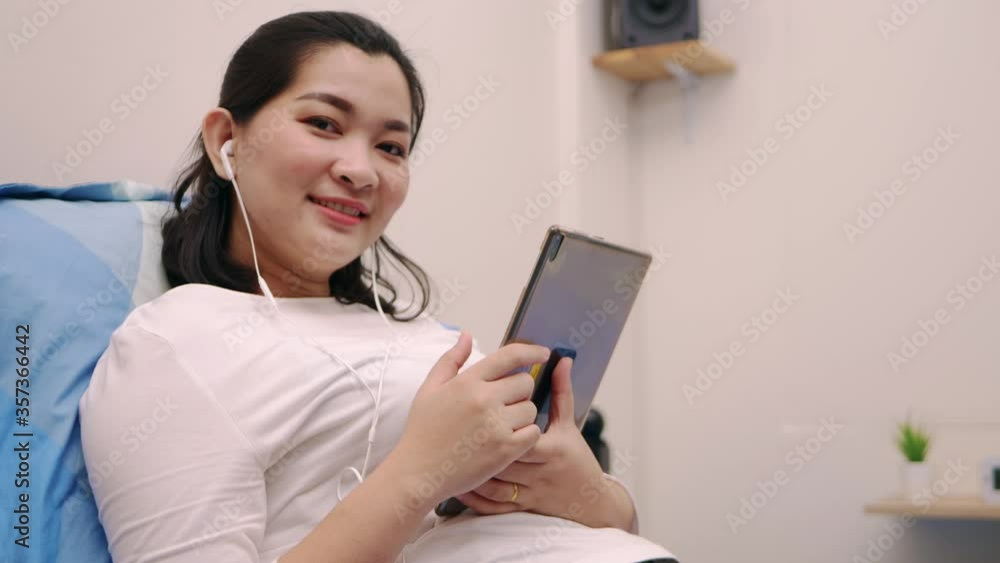 Beautiful asian woman look camera and smile relaxing and listening to music by tablet with headphones on the bed at home lifestyle and pleasure concept. Beautiful asian woman look camera and smile relaxing and listening to music by tablet with headphones on the bed at home lifestyle and pleasure concept.