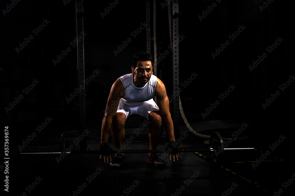 Selective focus of young Caucasian athlete postures working with barbell in a gym alone with dark background. A sport man workout in dark gym to build his body and strengths alone in shadow tone Selective focus of young Caucasian athlete postures working with barbell in a gym alone with dark background. A sport man workout in dark gym to build his body and strengths alone in shadow tone