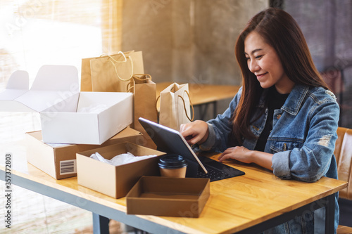 Closeup image of a young asian woman using tablet pc for online shopping with postal parcel box and shopping bags on the table at home