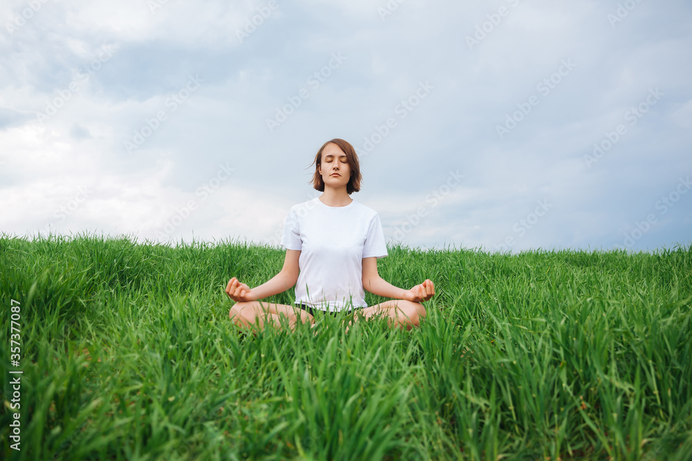 A young girl is meditating while sitting on the grass. Girl on the nature. Front view