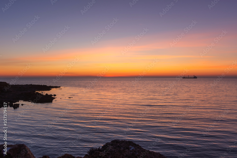Bright twilight sunset sky over the sea. Wonderful sky after sunset in orange shades. The natural background. Purple hue of the sea and sky. Abstract lines of fire clouds. A ship on the horizon.