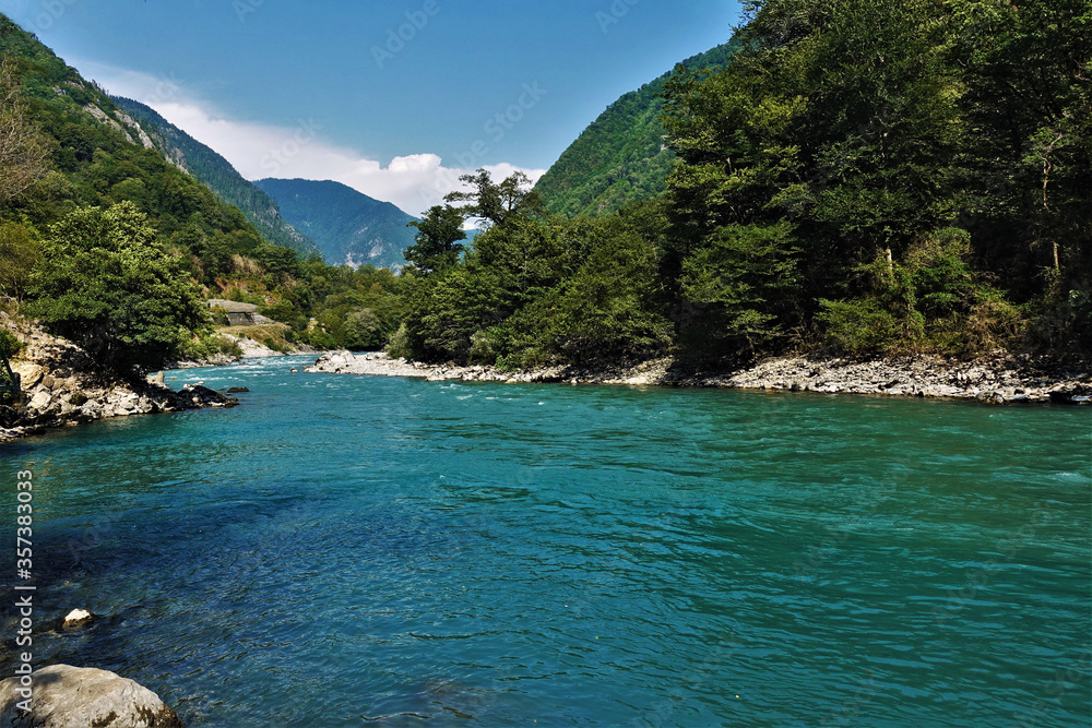 A river of aquamarine color flows between the slopes of the mountains covered with forest. The coast is rocky, the water is seething. Clouds on a bright blue sky. Abkhazia