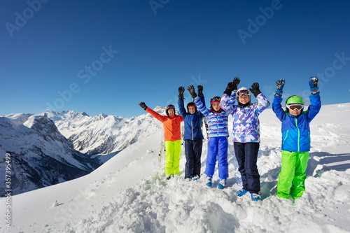 Big group of ski children stand on snow rise hands cheerfully smiling over mountain range peaks in colorful sport outfit