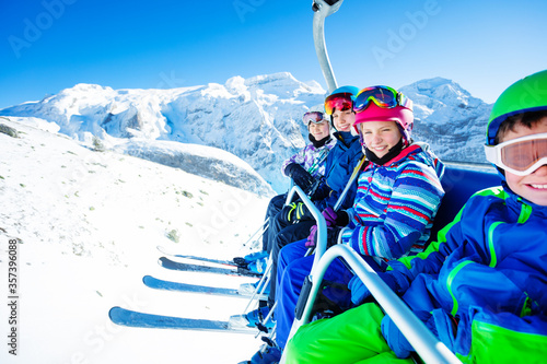 Group of children ski friends sit on chairlift lifting on the mountain top peak on sunny day