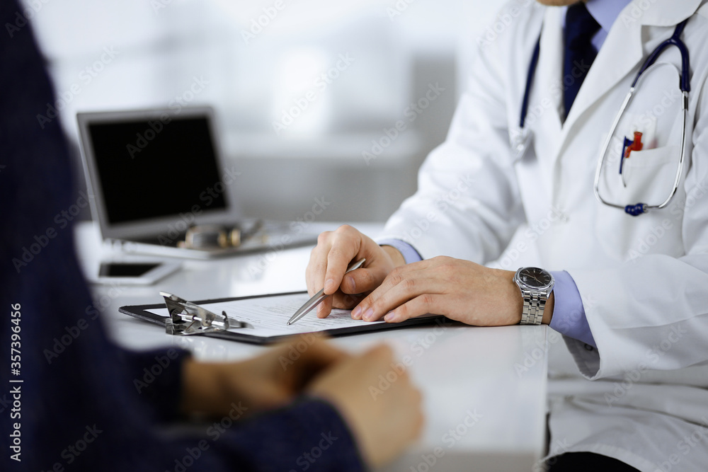 A doctor is talking to his patient, while sitting together at the desk in the cabinet in a hospital. Physician using clipboard for filling up medication history records. Perfect medical service in A doctor is talking to his patient, while sitting together at the desk in the cabinet in a hospital. Physician using clipboard for filling up medication history records. Perfect medical service in