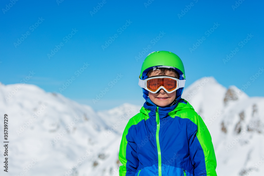 Close portrait of a boy in sport helmet and ski or snowboard glasses over bright sunny mountain snow tops on background