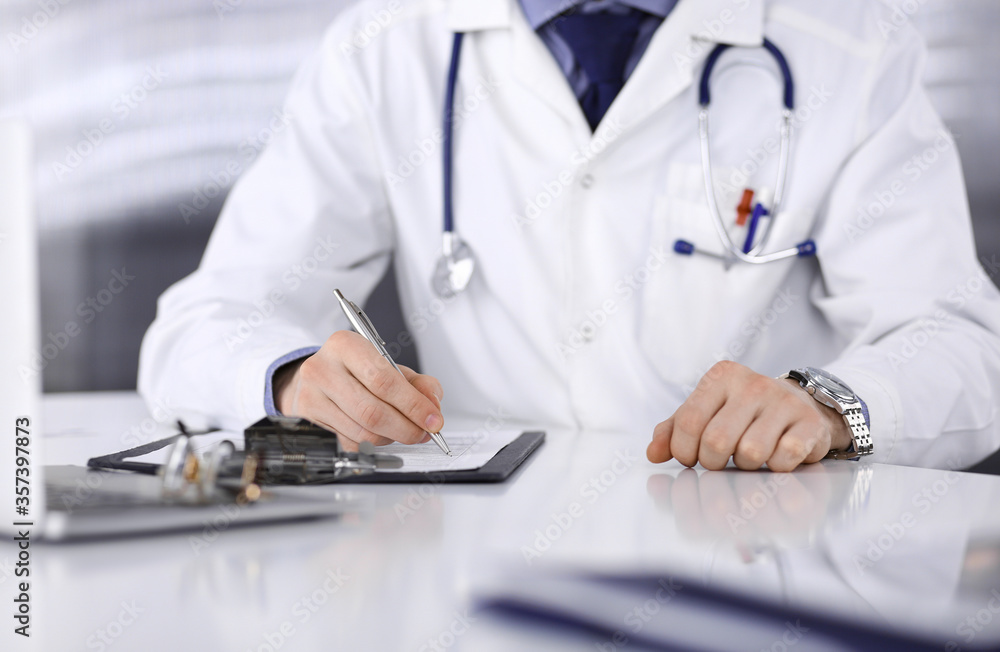 Unknown male doctor sitting and working with clipboard of medication history record in clinic at his working place, close-up. Young physician at work. Perfect medical service, medicine concept Unknown male doctor sitting and working with clipboard of medication history record in clinic at his working place, close-up. Young physician at work. Perfect medical service, medicine concept