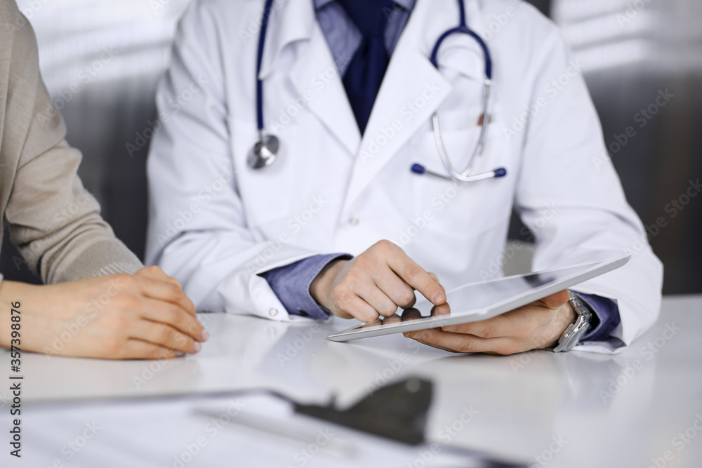 Unknown male doctor and patient woman discussing something while sitting in clinic and using tablet computer. Best medical service in hospital, medicine, pandemic stop Unknown male doctor and patient woman discussing something while sitting in clinic and using tablet computer. Best medical service in hospital, medicine, pandemic stop