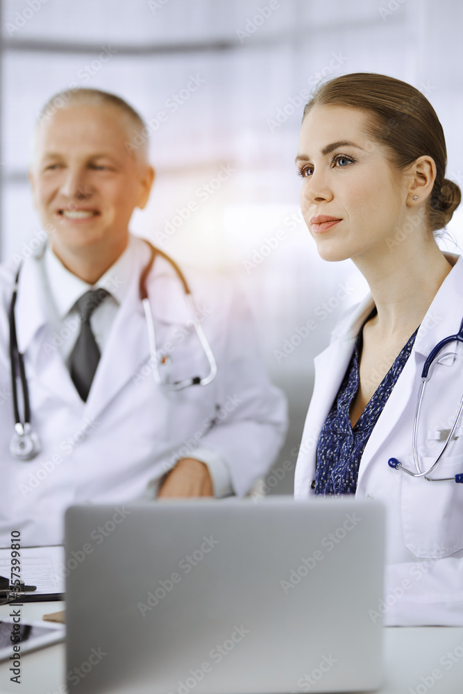 Elderly doctor and his female colleague discussing current disease therapy while sitting at working place in sunny clinic. Teamwork in medicine Elderly doctor and his female colleague discussing current disease therapy while sitting at working place in sunny clinic. Teamwork in medicine