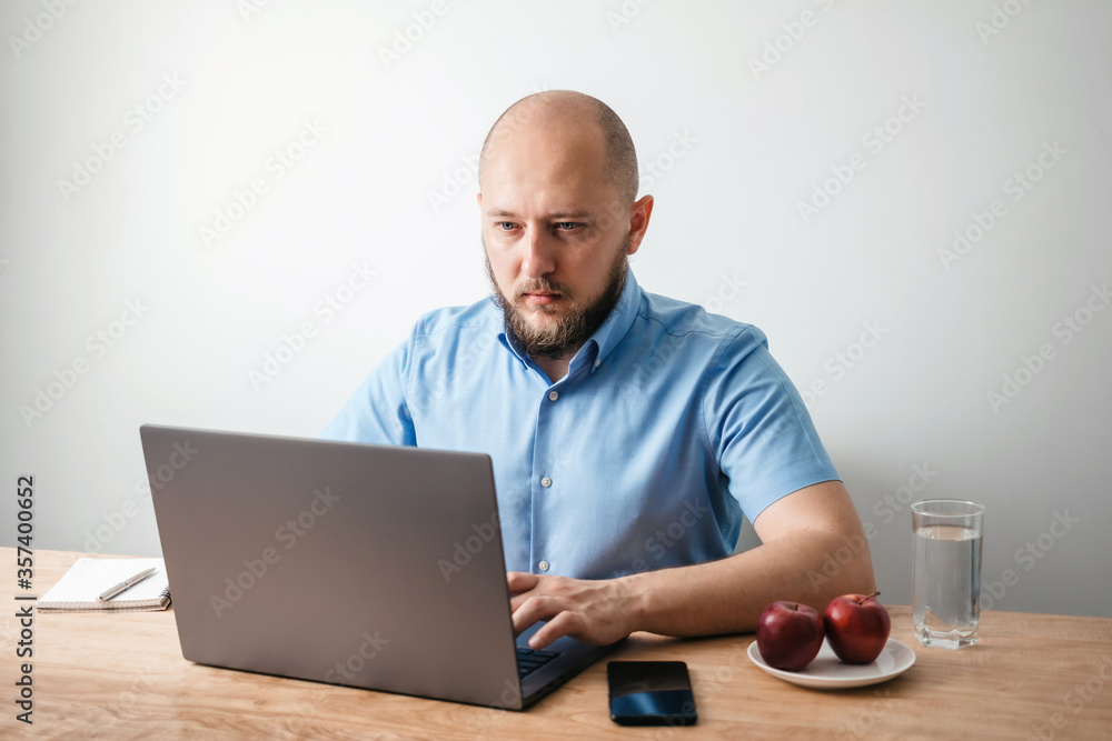 Bald young man with beard working online on his laptop at the wooden table in a home office with white wall as background. Business, freelance or study concept Bald young man with beard working online on his laptop at the wooden table in a home office with white wall as background. Business, freelance or study concept