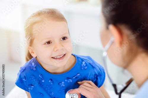 Doctor examining a child by stethoscope in sunny clinic. Happy smiling girl patient dressed in blue dress is at usual medical inspection