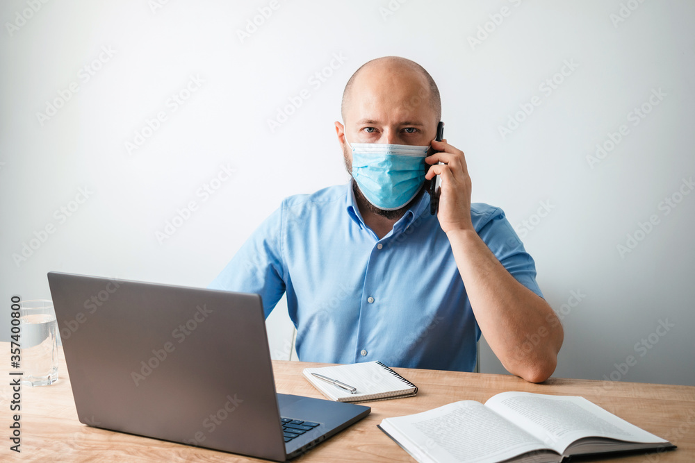 Bald business man with beard wearing surgical medical mask on face as protection against coronavirus, covid-19, talking on the phone, working or studying online on laptop at office Bald business man with beard wearing surgical medical mask on face as protection against coronavirus, covid-19, talking on the phone, working or studying online on laptop at office