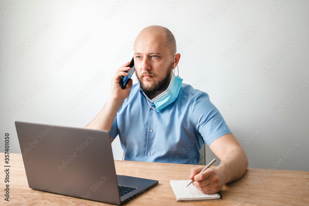 Bald business man with beard put off surgical medical mask as protection against coronavirus, covid-19 and talking on the phone, modern laptop on wooden table in office, white wall as background Bald business man with beard put off surgical medical mask as protection against coronavirus, covid-19 and talking on the phone, modern laptop on wooden table in office, white wall as background