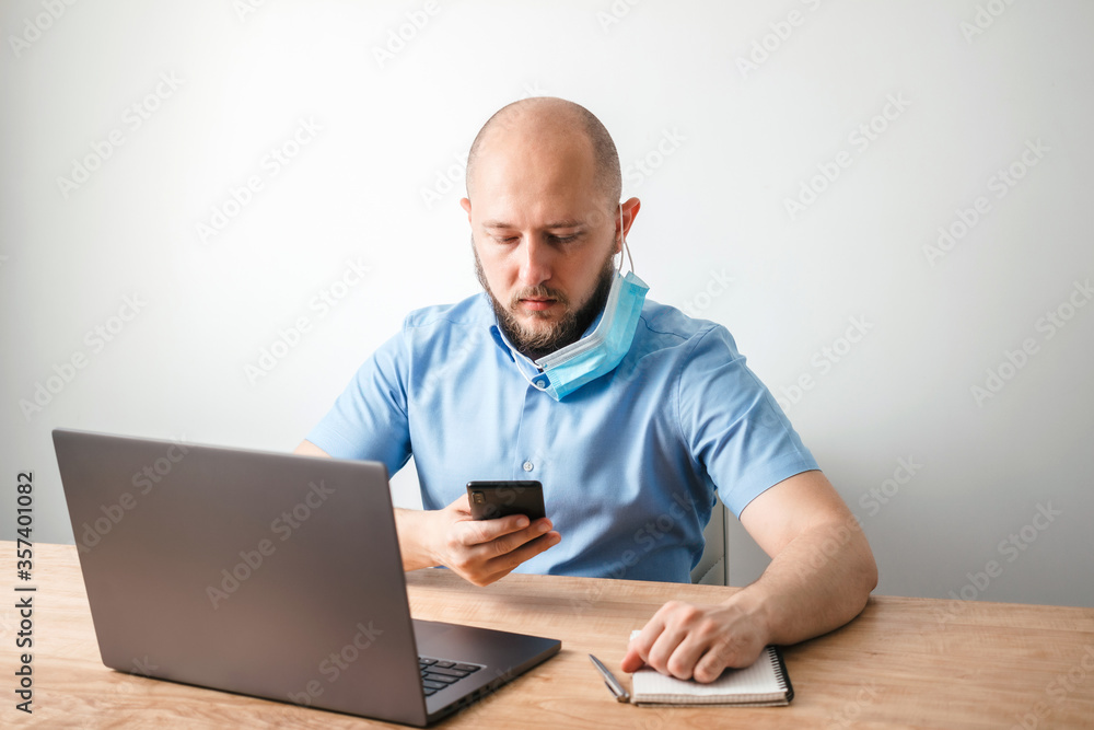 Bald business man with beard put off surgical medical mask as protection against coronavirus, covid-19 and uses smartphone, modern laptop on wooden table in home office, white wall as background Bald business man with beard put off surgical medical mask as protection against coronavirus, covid-19 and uses smartphone, modern laptop on wooden table in home office, white wall as background