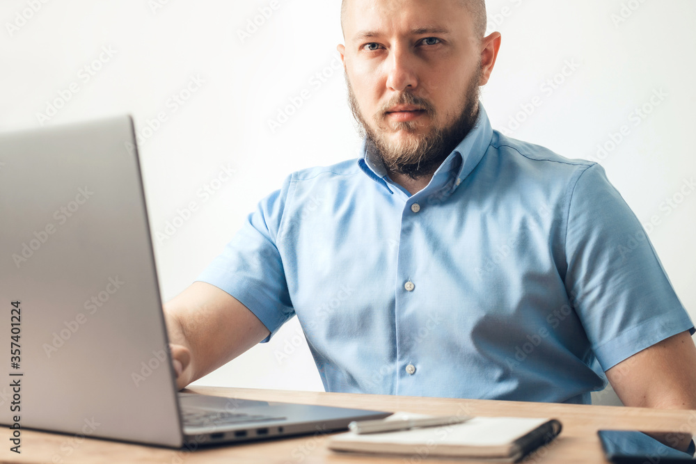 Bald young man with beard working online on his laptop at the wooden table in a home office with white wall as background. Business, freelance or study concept Bald young man with beard working online on his laptop at the wooden table in a home office with white wall as background. Business, freelance or study concept