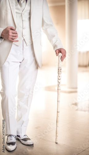 handsome elegant man in a white suit, shirts, vest, jacket, tie, stands in front of the camera, his right hand on the lapel of his jacket, his left hand on a wooden cane with a picture. 