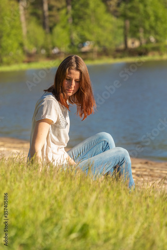 A red-haired girl sits in the grass by the lake and enjoys the evening sun during the sunset.