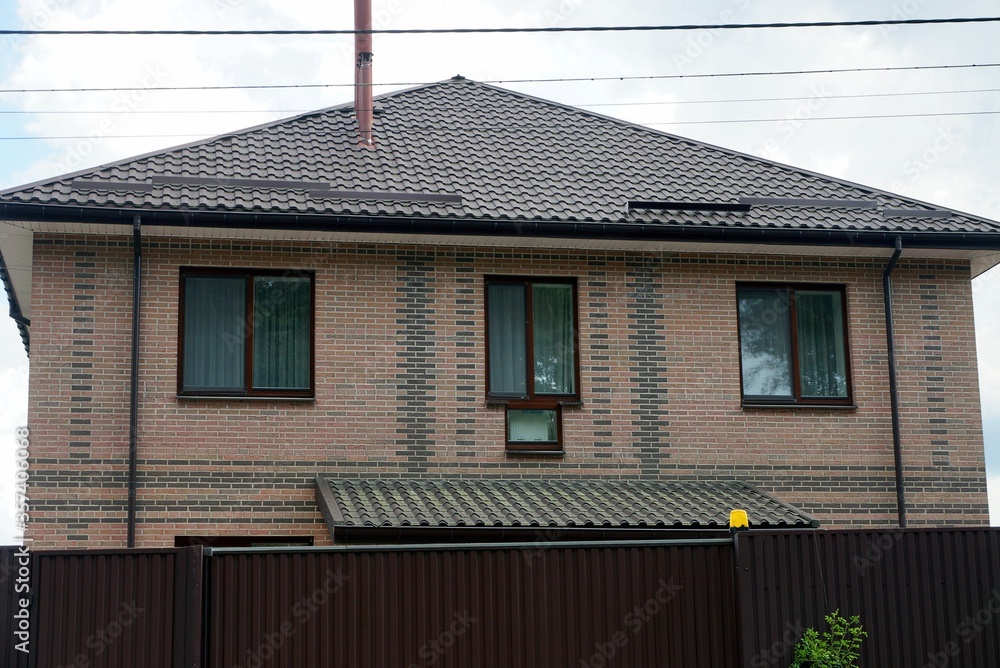 Naklejka premium facade of a brown private brick house with a row of windows under a tiled roof behind a metal fence against a gray sky