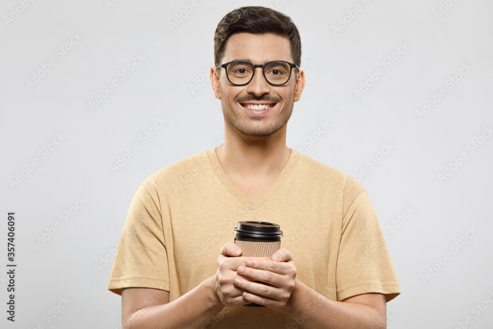 Young man in t-shirt and eyeglasses standing with takeaway coffee to go cup in hands, ready to enjoy his break from work, isolated on gray background Young man in t-shirt and eyeglasses standing with takeaway coffee to go cup in hands, ready to enjoy his break from work, isolated on gray background