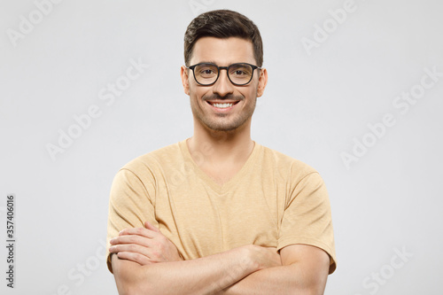 Young man in beige t-shirt and glasses, standing with arms crossed, smiling happily and feeling confident as professional, isolated on gray background