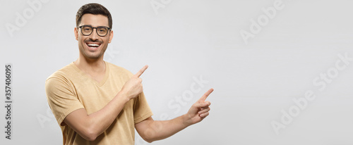 Banner of young smiling guy in beige t-shirt and eyewear, showing commercial offer on right, pointing to it with both hands, isolated on gray background photo