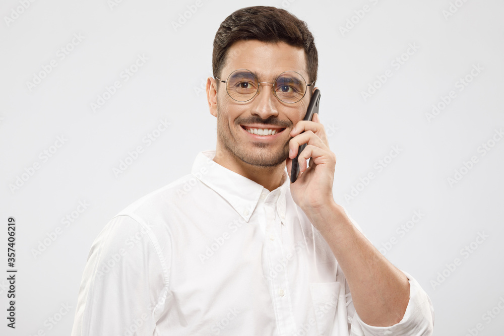 Portrait of young positive handsome businessman in shirt and glasses, answering phone call, looking aside with smile, isolated on gray background Portrait of young positive handsome businessman in shirt and glasses, answering phone call, looking aside with smile, isolated on gray background