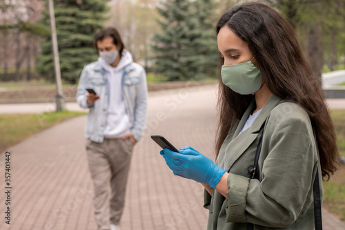 Young woman in green mask and gloves standing in city park and texting sms on smartphone, social distancing during coronavirus concept