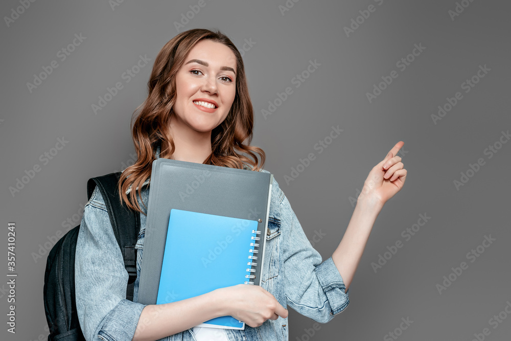 Student girl holds folders, books, notebooks, notebooks in hands and points a finger at copy space isolated on gray wall background Student girl holds folders, books, notebooks, notebooks in hands and points a finger at copy space isolated on gray wall background