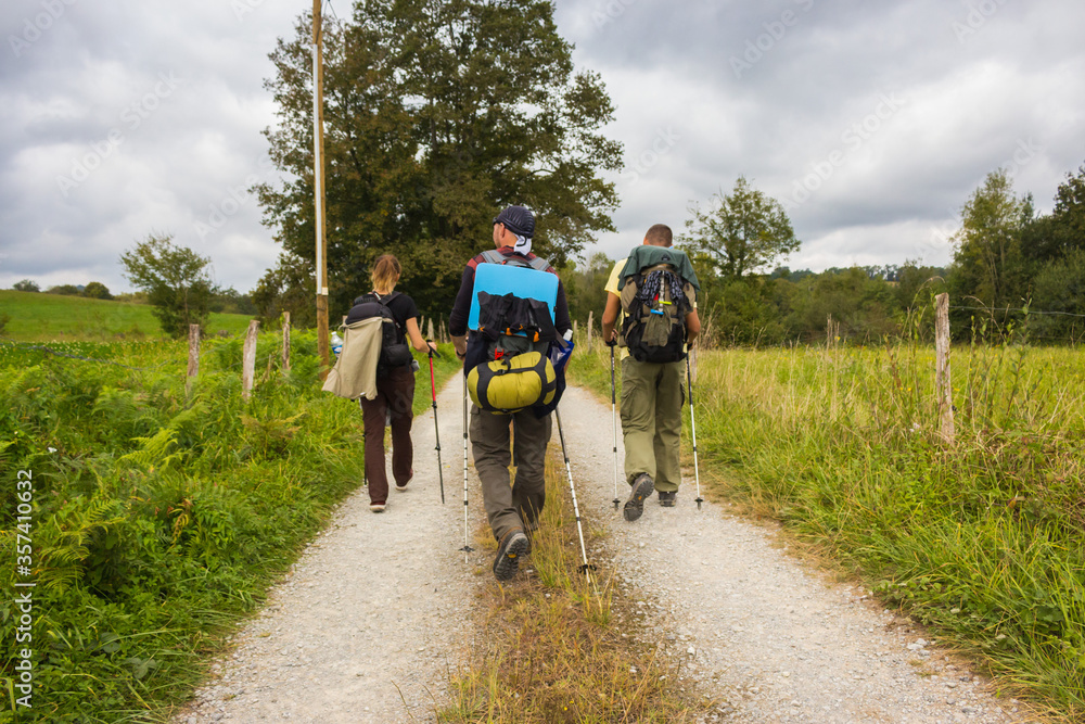 Fototapeta premium Spain - 10/01/2019: Three unknown pilgrims walking on Camino de Santiago. Group of tourists with backpacks on village road. Active people on the way. Pilgrimage concept. Backpackers from the back.