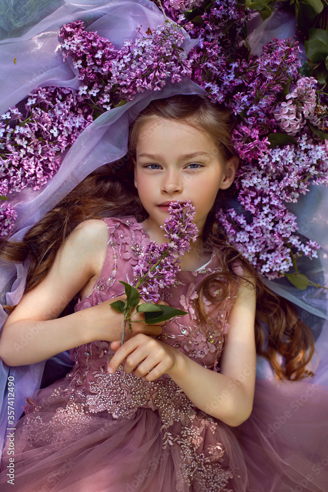 Fototapeta premium child girl in a purple floral dress lies on the ground among lilacs