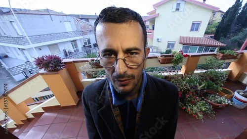 Uomo con giacca e occhiali da vista bussa insistentemente  ad una porta  stando fuori nel terrazzo adiacente, sullo sfondo altre abitazioni in lontananza 