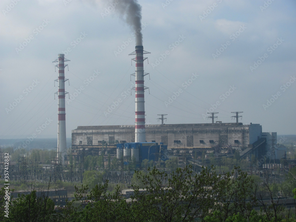 There are green bushes near, and in the distance there is grey smoke coming out of the pipe. The borderline of nature and industrial landscape.