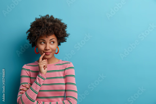 Dreamy beautiful woman thinks about something pleasant  makes plans for future  keeps finger near lips  concentrated aside  wears casual striped jumper  isolated on blue background copy space for text