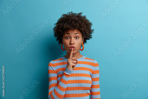 Shush, be quiet! Mysterious dark skinned woman makes hush sign, presses finger to lips, demands silence and secrecy, wears striped sweater, asks to keep secret, wears casual sweater, isolated on blue