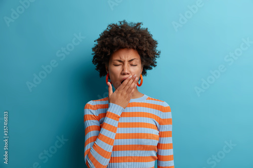 Tired exhausted Afro American woman feels bad after hard day at work, covers mouth and has sleepy expression, wears casual striped jumper, isolated on blue background. Restless and sleepiness concept
