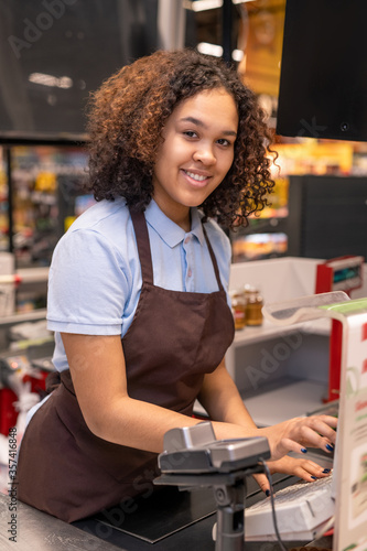 Obraz na płótnie Happy young mixed-race cashier with dark wavy hair pressing button on cashbox