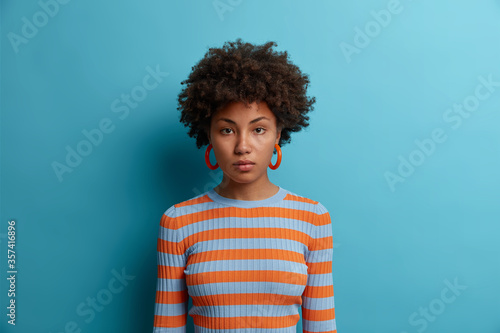 Portrait of serious girlfriend with calm confident attentive expression, wears casual striped sweater, poses indoor against blue background, stands with hands down. Determined female freelancer