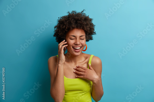 Carefree cheerful dark skinned woman has funny amusing conversation via phone, discusses something joyfully with best friend and laughs, hears funny joke, wears summer clothes, isolated on blue