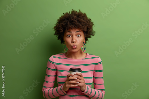 Lovely dark skinned woman with Afro hairdo drinks takeaway coffee, holds paper cup, keeps lips rounded, dressed in casual striped jumper, has coffee break after work, isolated on green studio wall © Wayhome Studio