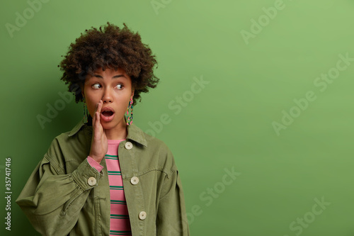 Surprised dark skinned millennial woman gossips and tells secret, keeps mouth opened, looks aside, doesnt want someone hear her story, wears fashionable clothing, isolated on green studio background