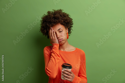 Sleepy exhausted dark skinned curly woman holds cup of coffee, cannot wake up and go to work, had sleepless night, wears casual orange jumper, has sad expression, isolated on green background