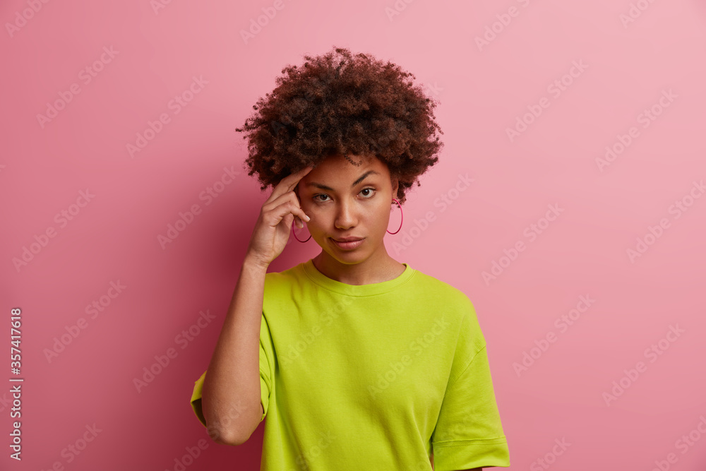 Photo of serious dark skinned woman keeps hand on head, tries to remember something, makes decision and looks thoughtfully, wears bright summer clothes, isolated on rosy background. Face expressions Photo of serious dark skinned woman keeps hand on head, tries to remember something, makes decision and looks thoughtfully, wears bright summer clothes, isolated on rosy background. Face expressions