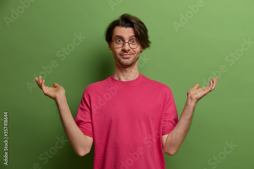 Clueless hesitant young man raises hands in no clue, tells who cares, cannot decide, has confused expression, wears transparent glasses and pink t shirt, shrugs shoulders, isolated on green background