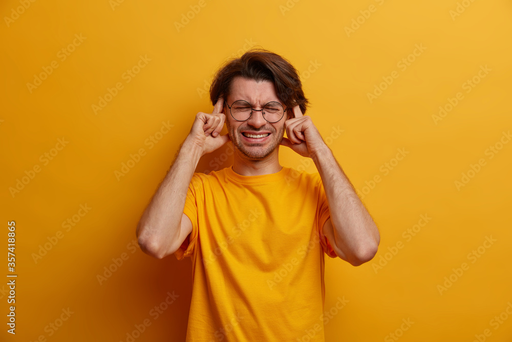 Distressed man squints face, touches temples, has painful feelings in head, suffers from migraine, clenches teeth to reveal pain, closes eyes, wears bright yellow t shirt in one tone with background Distressed man squints face, touches temples, has painful feelings in head, suffers from migraine, clenches teeth to reveal pain, closes eyes, wears bright yellow t shirt in one tone with background