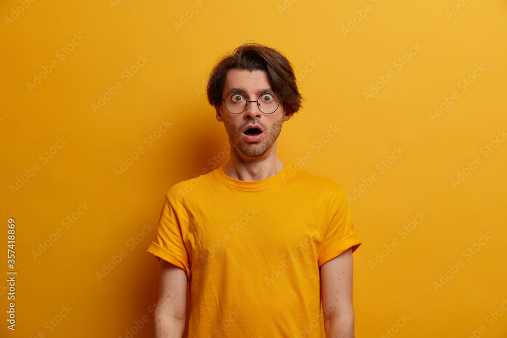 Portrait of astonished brunet man gasps from great wonder, keeps mouth widely opened, reacts on shocking news, stands speechless, wears optical glasses and t shirt, yellow background. Monochrome Portrait of astonished brunet man gasps from great wonder, keeps mouth widely opened, reacts on shocking news, stands speechless, wears optical glasses and t shirt, yellow background. Monochrome