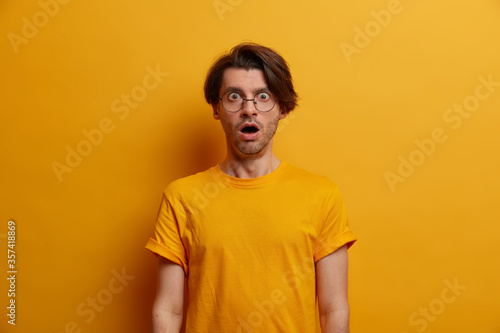 Portrait of astonished brunet man gasps from great wonder, keeps mouth widely opened, reacts on shocking news, stands speechless, wears optical glasses and t shirt, yellow background. Monochrome © Wayhome Studio